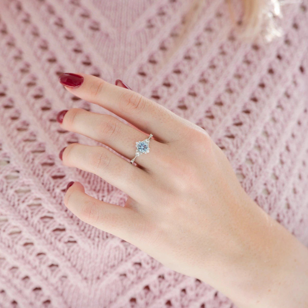Silver ring with blue center and clear halo worn on a hand against pink knitwear